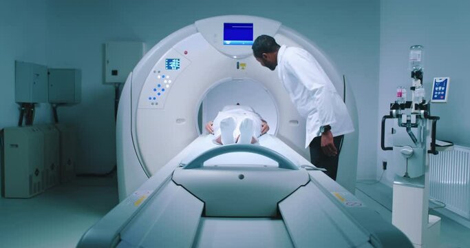Patient is undergoing MRI procedure. Multucultural medical worker performs magnetic resonance imaging. Patient is lying at MRI capsule. Front view of modern equipped tomography room.