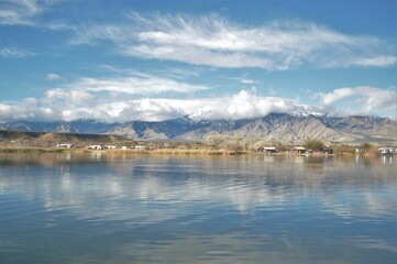 Beautiful natural landscape of high mountains next to farmlands near blue lake reflecting cloudy sky