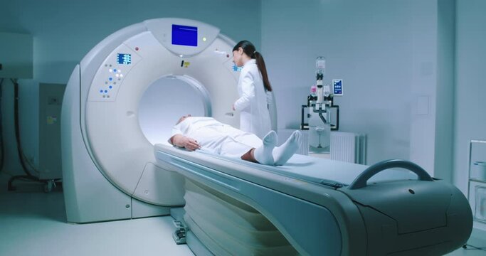 Female medical worker dressed up in gown before MRI examination. Female doctor is preparing patient for magnetic resonance procedure. Patient is lying at CT scanner bad and waiting to be scanned.
