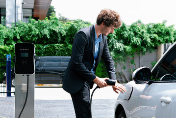 Progressive businessman with electric car recharging at public charging station at modern city...