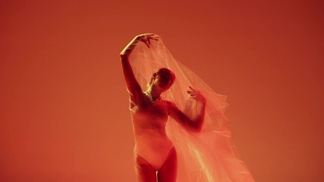 Lightness. Tender young woman, ballerina dancing with transparent fabric against orange studio background in neon light. Concept of classical art, dance, beauty, inspiration, creativity