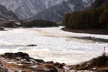 A turn of the bed of a slightly melted river flowing on a winter evening at the foot of a snow-covered mountain range.