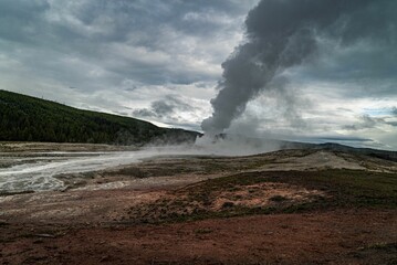 Fototapeta na wymiar Scenic view of white smoke from a geyser with green mountain in the background on a cloudy day
