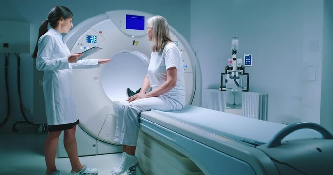 Zoom shooting. Female doctor talking to patient and points to MRI tomography scanner.. Female patient is sitting at CT scanner bed and listening to doctor. Medical worker holds tablet and scrolles it.
