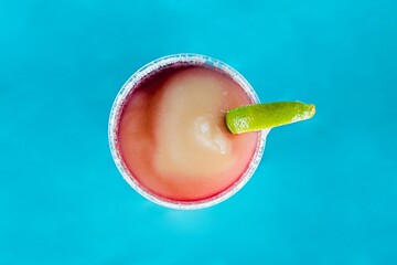 Top view of a round glass with an iced cocktail and a lime slice on a blue surface