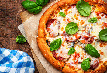 Pizza with spicy salami sausage, mozzarella cheese, tomato sauce and green basil on rustic wooden kitchen table background, top view