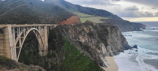 View of a bridge spanning across a lush green hillside overlooking the crashing waves of the ocean