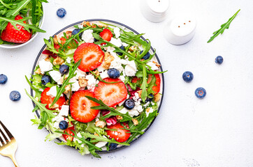 Strawberry salad with arugula, lettuce, blueberries, feta cheese and walnuts, white table. Fresh...