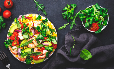 Fresh salad with shrimps, sweet corn, avocado, red tomatoes, lamb lettuce and onion on black table background. Top view