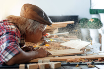 Asian senior carpenters working in the workshop, blowing dust off of a piece of wood, concept of  works in a carpentry shop