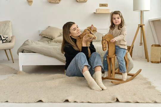A little child rides on a wooden horse next to her mom who hugs a cat