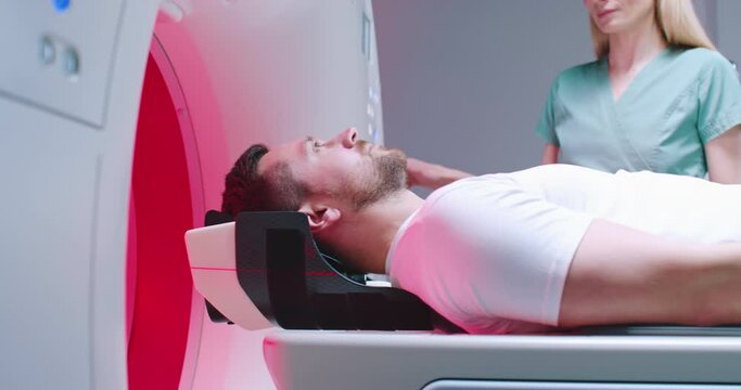 Shoot from side. Male patient is undergoing MRI tomography examination. Female doctor dressed up in blue gown is adjusting CT scanner. Man is moving into MRI capsule. Tomography under infrared rays.