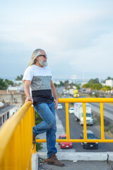 Long-haired gray-haired man wearing dark glasses on a pedestrian bridge. Cityscape in the background.