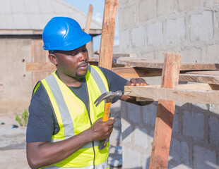 A happy male African construction worker with a blue safety helmet on his head, working with an hammer on a building site in Nigeria