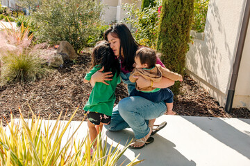Mother with infant son hugging her toddler daughter in front of house