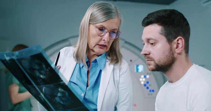 Close-up of patient and doctor are having discussion. Female doctor is showing tomography scans to patient. Middle-aged doctor is talking to male patient. Woman doctor in glasses explain MRI results.