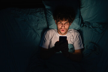 Shocked tired man with insomnia watching video on cellphone while lying on bed in dim bedroom at night at home. Can not sleep. Insomnia, addiction concept.