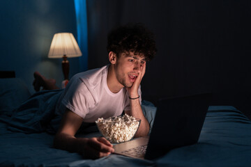 Shocked young male eating popcorn and watching film on laptop while resting on bed at night at home