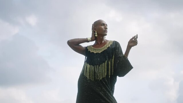 Gay bipoc man in luxury dress, boutique brass jewelry, makeup poses gracefully tropical rocky hill top on ocean beach. Homosexual ethnic elegant person standing on stone like Oshun orisha. Cloudy sky.