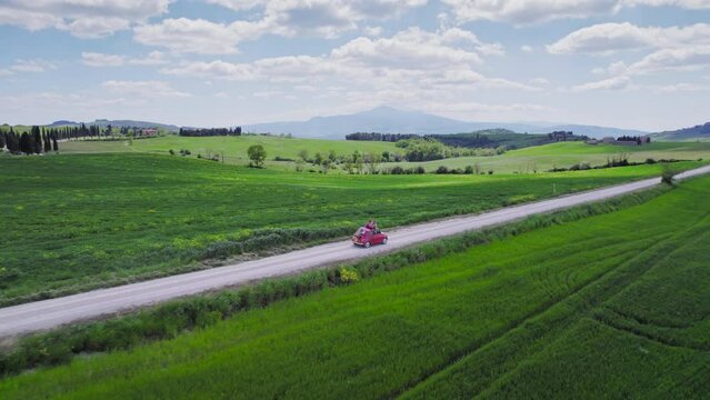 Aerial tracking shot of red car driving by green Tuscany landscape
