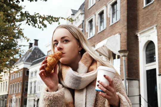 Young blond woman with croissant drinking coffee on a street