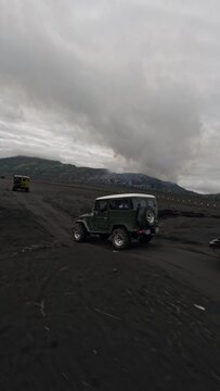 Vertical video. Yellow retro colorful jeep SUV riding black volcano lava field dirt dust desert at mountain aerial view. FPV drone shot speed automobile rally safari travel extreme sport auto drive 4k