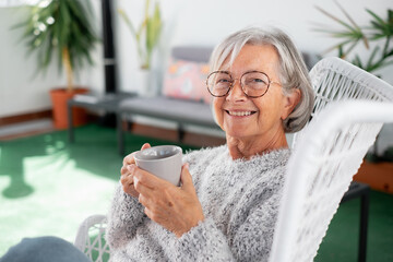 Portrait of senior attractive woman with cheerful smile relaxing at the armchair in the balcony at home looking at camera holding a coffee or tea cup