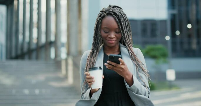 Happy corporate Black woman walking with coffee-to-go and using her smartphone. 