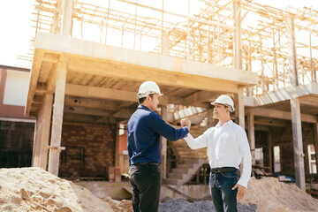 2 Asian male engineers, mechanic manager Holding hands together about working with energy and unity Wearing uniforms, helmets, standing on a construction site about a large industrial architecture.