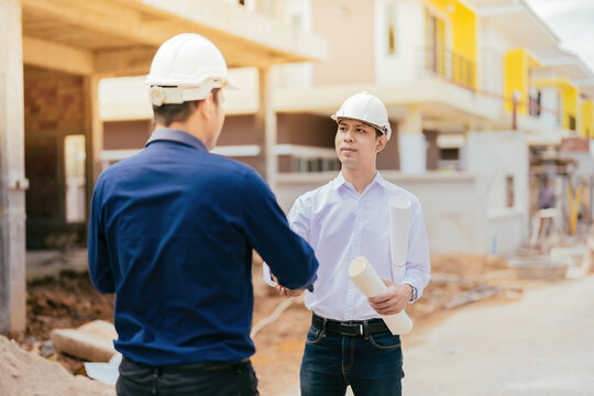 Half-body shot over the shoulder between two Asian male engineers holding hands. in the design of construction work in the architectural industry holding a blueprint wear a helmet talking about design