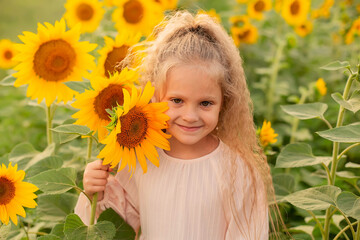 a blonde girl with long hair in a pink linen dress stands in a field with sunflowers and holds a flower in her hands