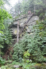 A steep rocky wall made of naturally cracked granite rocks covered with green moss, some shrubs, tall tree trunks, a broken tree trunk in the Polish part of The Karkonosze Mountains, Sudetes