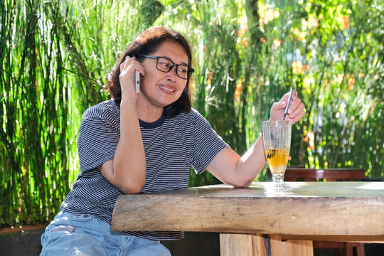 A middle-aged woman is talking on the phone while stirring her iced tea
