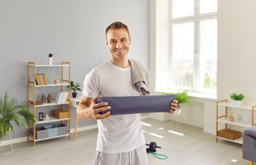 Portrait of a happy man with a towel on his shoulder holding a yoga mat, looking at camera and smiling. Sport, workout, fitness exercise, wellness, physical activity at home concept