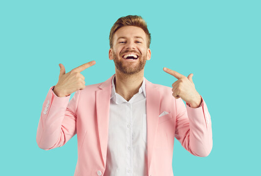Portrait of a young handsome bearded man wearing bright pink suit and white shirt pointing at himself with index finger smiling positive and cheerful at camera isolated on studio blue background.