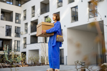 Young woman carrying cardboard box and flowerpot at inner yard of apartment building, relocating to a new home