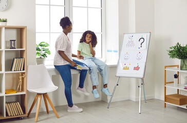 Little school child and counseling psychologist talking about emotions while sitting on window sill next to office whiteboard with pictures of emoji. Therapy, psychology, communication concept