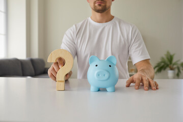 Cropped shot of man sitting at white desk with blue piggy bank and wooden question mark...
