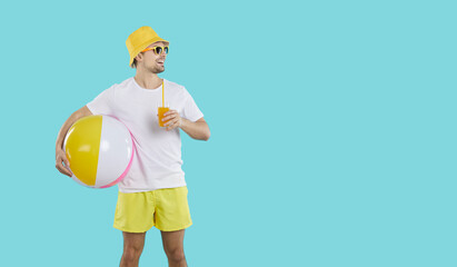 Man enjoying summer holiday. Happy guy wearing white tee shirt, vacation shorts, yellow panama hat and sun glasses standing on blue studio background, holding beach ball and looking at copy space side