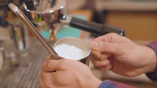 Preparing steamed milk in stainless steel milk jug. Closeup of male barista hands