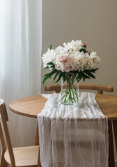 A bouquet of beautiful white peonies in a glass vase on a wooden round table with a white tablecloth