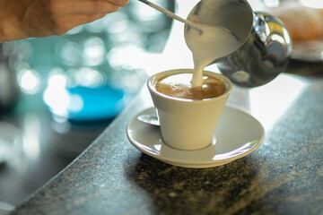 Hands of young Woman barista pouring steamed creamy milk on cappuccino cup at the bar counter.