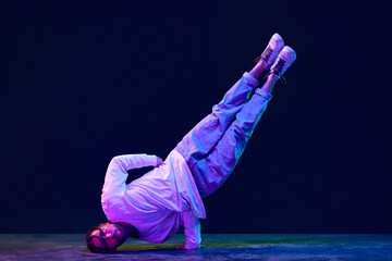 Young guy in sport style clothes dancing breakdance, hip-hop against black studio background in neon light. Strong body