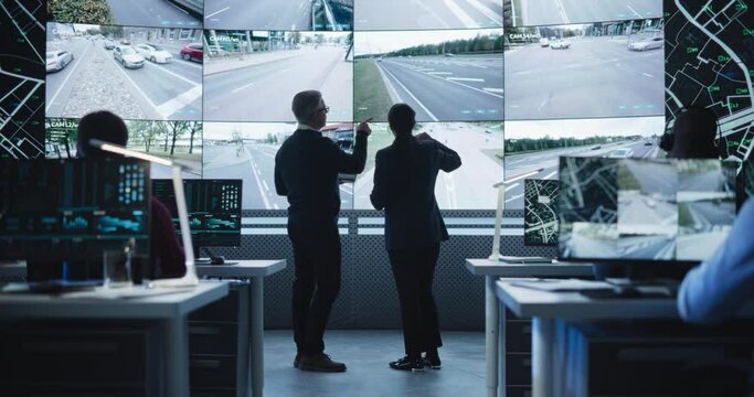 Traffic Commission Project Manager and Road Planning Department Computer Science Engineer Talking while Using Big Digital Display in a Monitoring Room. Screen Showing a Cluster of Live Traffic Feeds