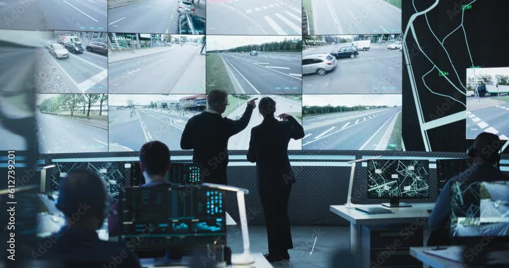 Poster Two Multiethnic Software Engineers Talking Next to a Big Screen in a Modern Monitoring Office with Live CCTV Footage with Traffic Situation. Monitoring Room Big Data Engineers Work on Computers