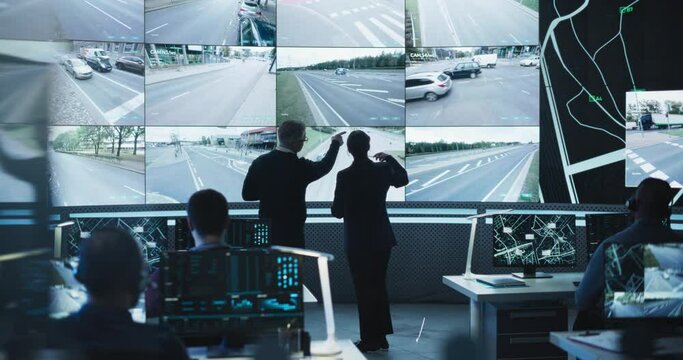 Two Multiethnic Software Engineers Talking Next to a Big Screen in a Modern Monitoring Office with Live CCTV Footage with Traffic Situation. Monitoring Room Big Data Engineers Work on Computers