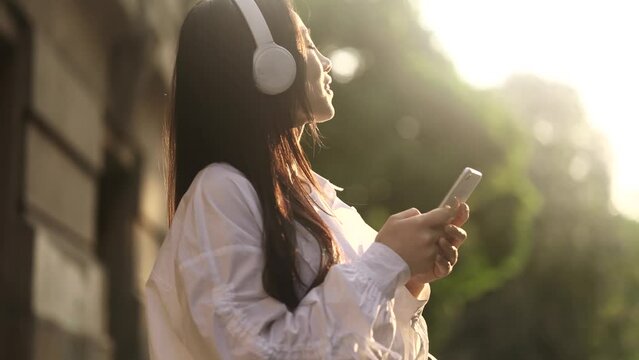 Charming asian woman with headphones hold smartphone scrolling browsing online and listening to music at urban city Pretty female relax enjoying great day with beautiful sun rays on her face outdoors