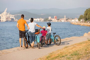 Two kids, with helmets on their heads, and smiling parents riding bikes on a family-friendly cycle route along a sea coastline, front view.