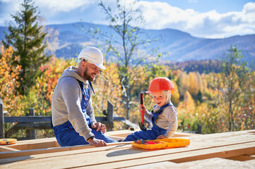 Father with toddler son building wooden frame house. Male builders hammering nail into plank on...