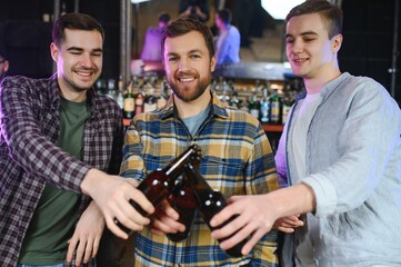 Three young men in casual clothes are smiling and clanging glasses of beer together while sitting at bar counter in pub